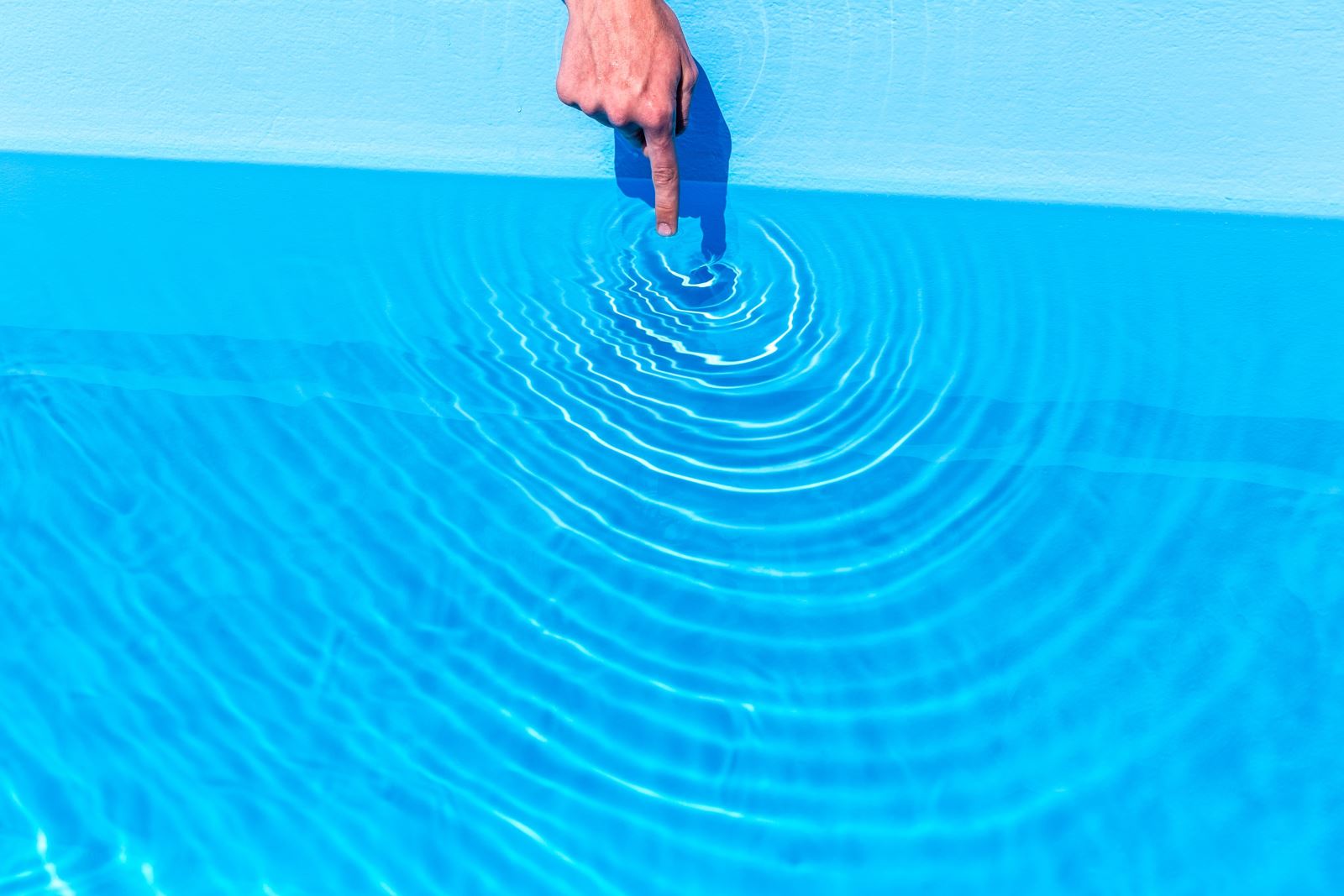 A hand stretching into a swimming pool and touching the water, causing a rippling effect